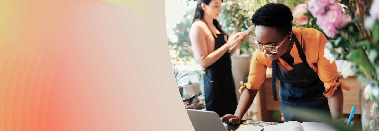 Two individuals wearing aprons are working in a flower shop. One person is leaning over a counter, looking at a laptop, while the other is standing in the background, holding a phone.