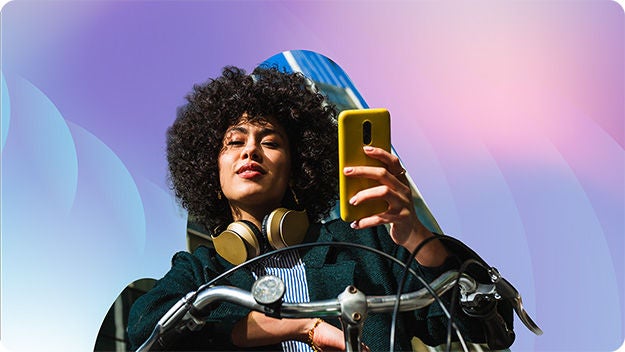 A person with curly hair, wearing headphones around the neck, is holding a yellow smartphone.