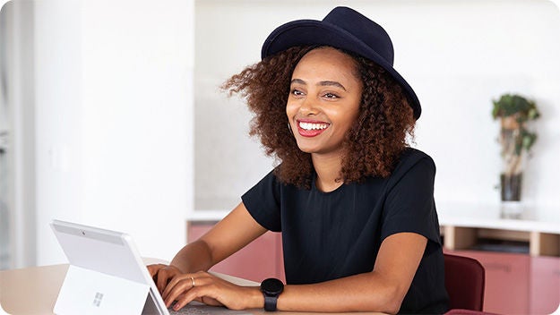 A person with curly hair is sitting at a desk, typing on a laptop. The person is wearing a black shirt and a dark hat. The background shows an indoor setting with some furniture and a plant in the distance.