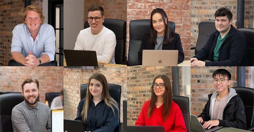  A group of seven people sitting at desks with laptops in an office setting. The background features exposed brick walls and large windows.