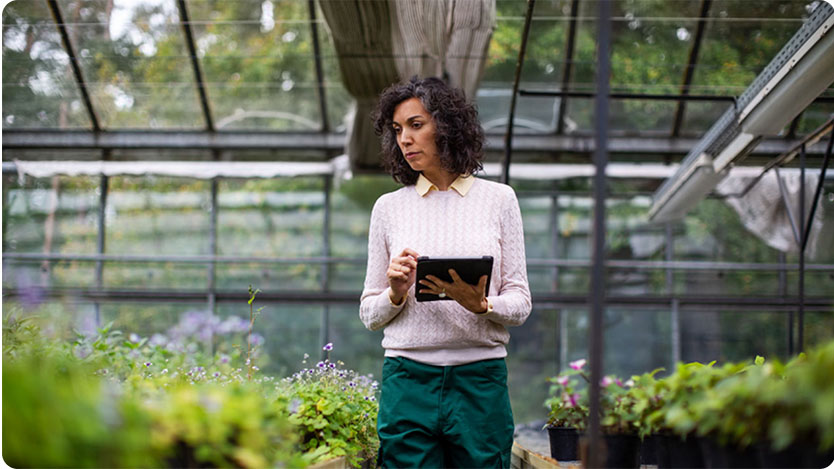 A person is standing in a greenhouse, holding and using a tablet. The greenhouse is filled with various plants on both sides of the person, who is dressed in a light sweater and green pants.
