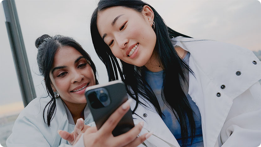 Two individuals in light-colored jackets looking at a smartphone together outdoors under a cloudy sky.