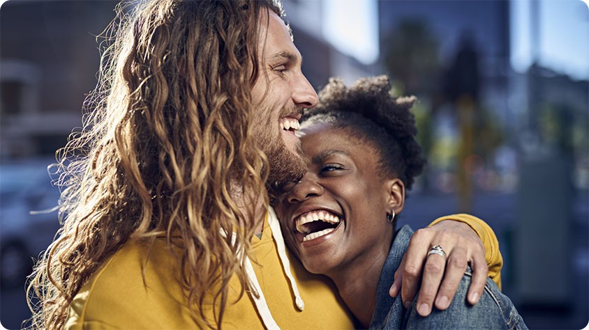 Two people standing close together outdoors in an urban setting. One has long, wavy hair and wears a yellow hoodie with an arm around the other, who has short, curly hair and wears a denim jacket. Blurred buildings and trees are visible in the background.
