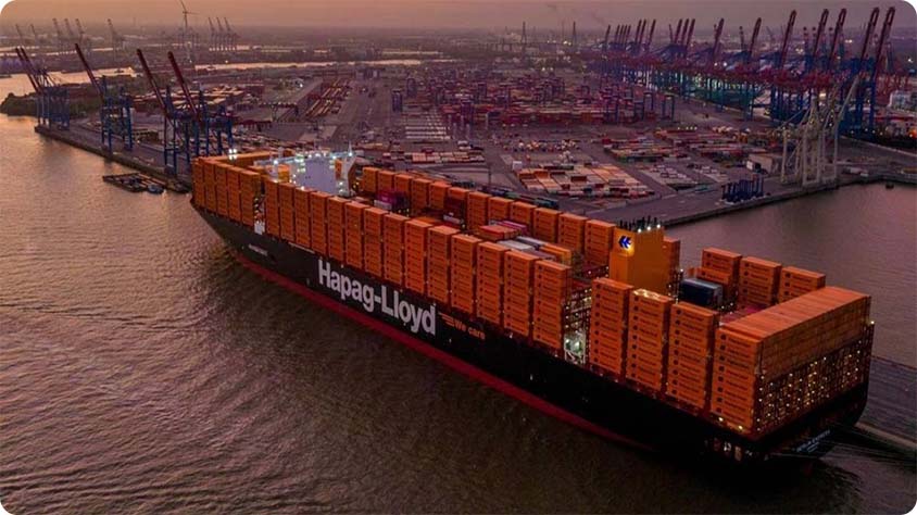 A large cargo ship labeled "Hapag-Lloyd" is docked at a busy port during sunset. The ship is loaded with numerous orange shipping containers.