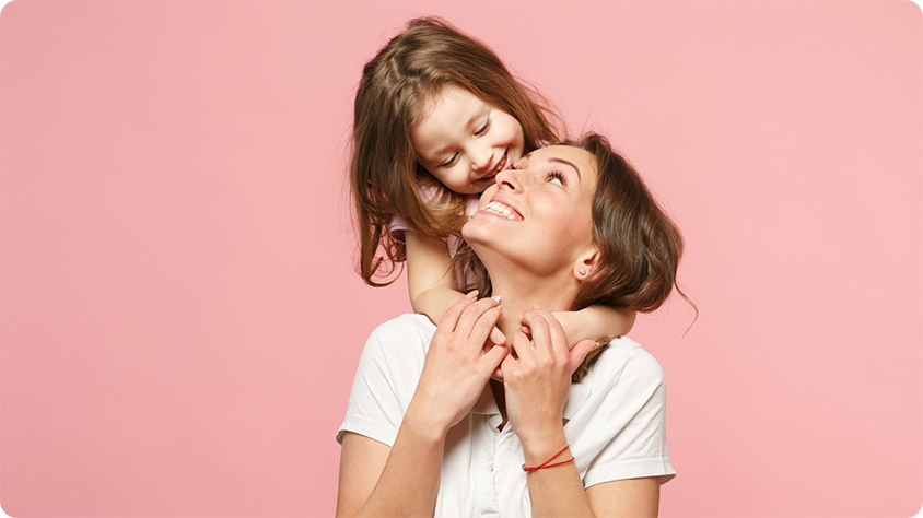 A person wearing a white shirt is holding a child who is hugging them from behind. The background is a solid pink color.