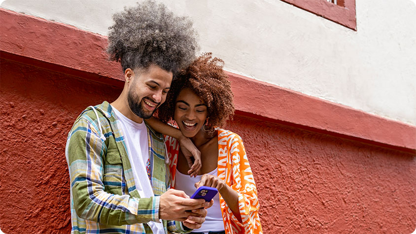 Two people with curly hair stand close together against a red and white textured wall, smiling as they look at a smartphone.