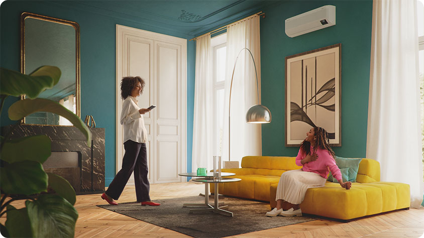 A modern living room with teal walls and large windows covered by sheer white curtains. The room features a yellow sectional sofa, a round glass coffee table, and a tall floor lamp with a metallic shade. Two people are present; one is sitting on the sofa wearing a pink top and white skirt, while the other is standing near the door holding a smartphone.