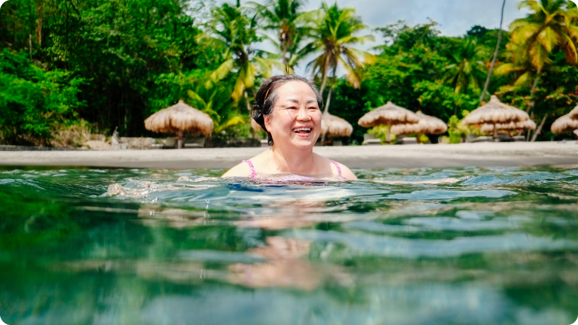 A person is swimming in clear, greenish-blue water near a tropical beach. The background features several thatched-roof huts and tall palm trees, with lush greenery and a partly cloudy sky.